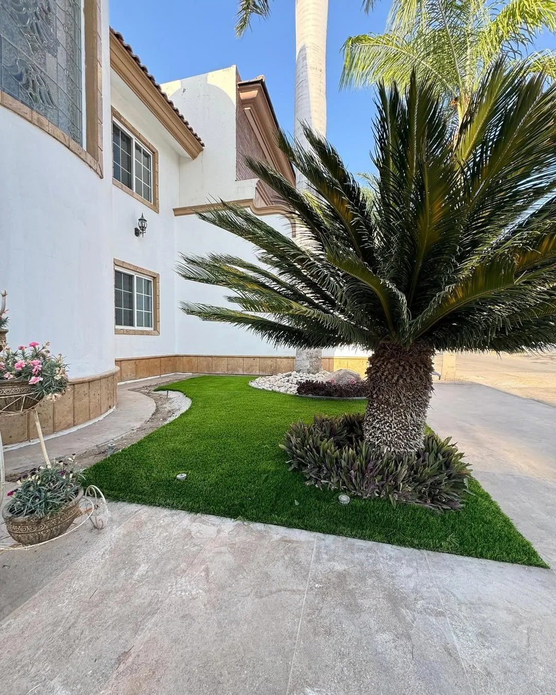 Jardín con césped verde, planta de palma en maceta, casa blanca de paredes con ventanas y paredes decorativas en estilo mediterráneo, cielo azul, y fondo con palmeras.