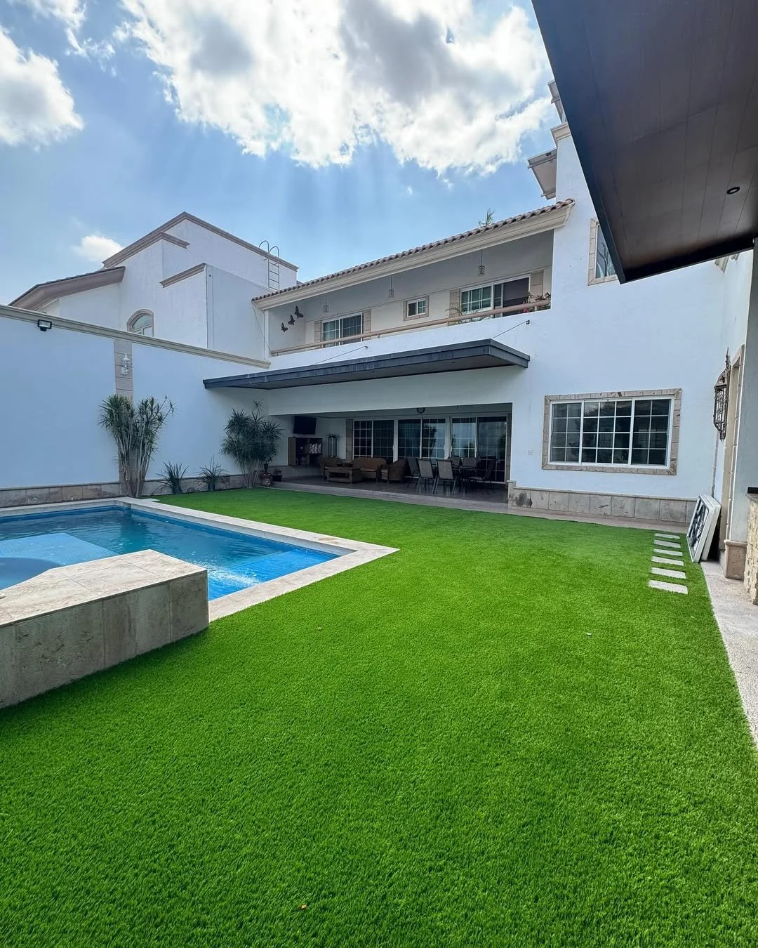 Patio trasero con césped artificial, piscina y muebles de exterior en una casa de dos pisos blanca, con cielo nublado y rayos de sol