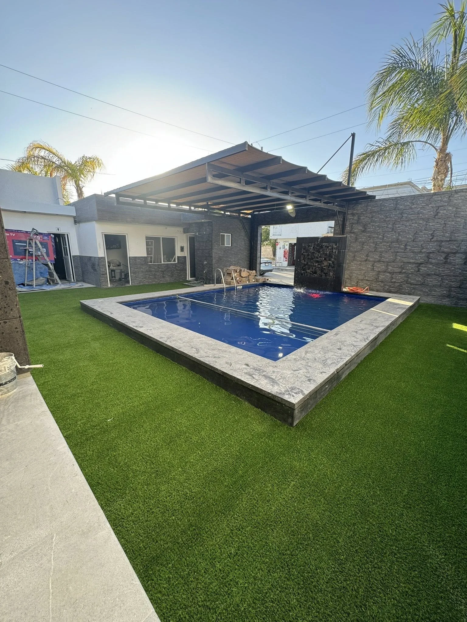 Piscina rectangular rodeada de césped artificial y una pared de piedra. Tiene una cubierta de madera con techo, y en una esquina hay una cascada o fuente de agua. Al fondo, un edificio y palmeras.