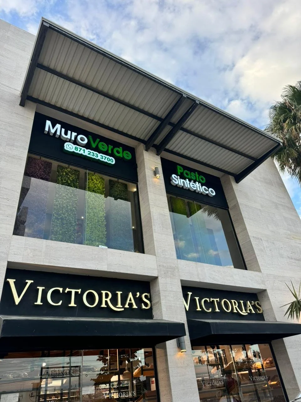 Fachada de un edificio con letrero de 'Victoria's' en negro y letras doradas, y signos de negocios como 'Muro Verde' y 'Pasta Sintetico' en la parte superior, con ventanales grandes y cielo parcialmente nublado.