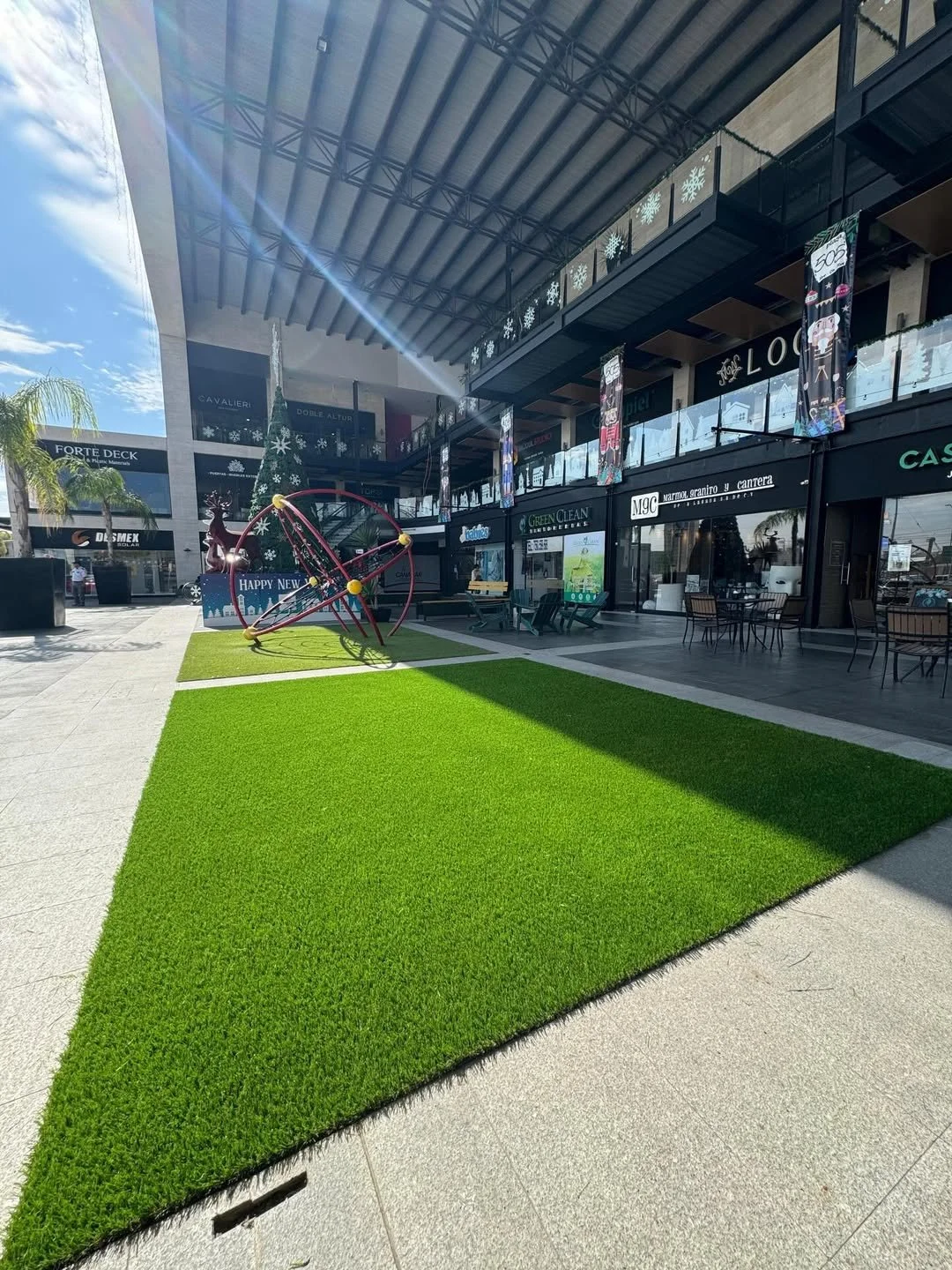 Plot de césped artificial decorativo en la entrada de un centro comercial con juegos infantiles y tiendas.