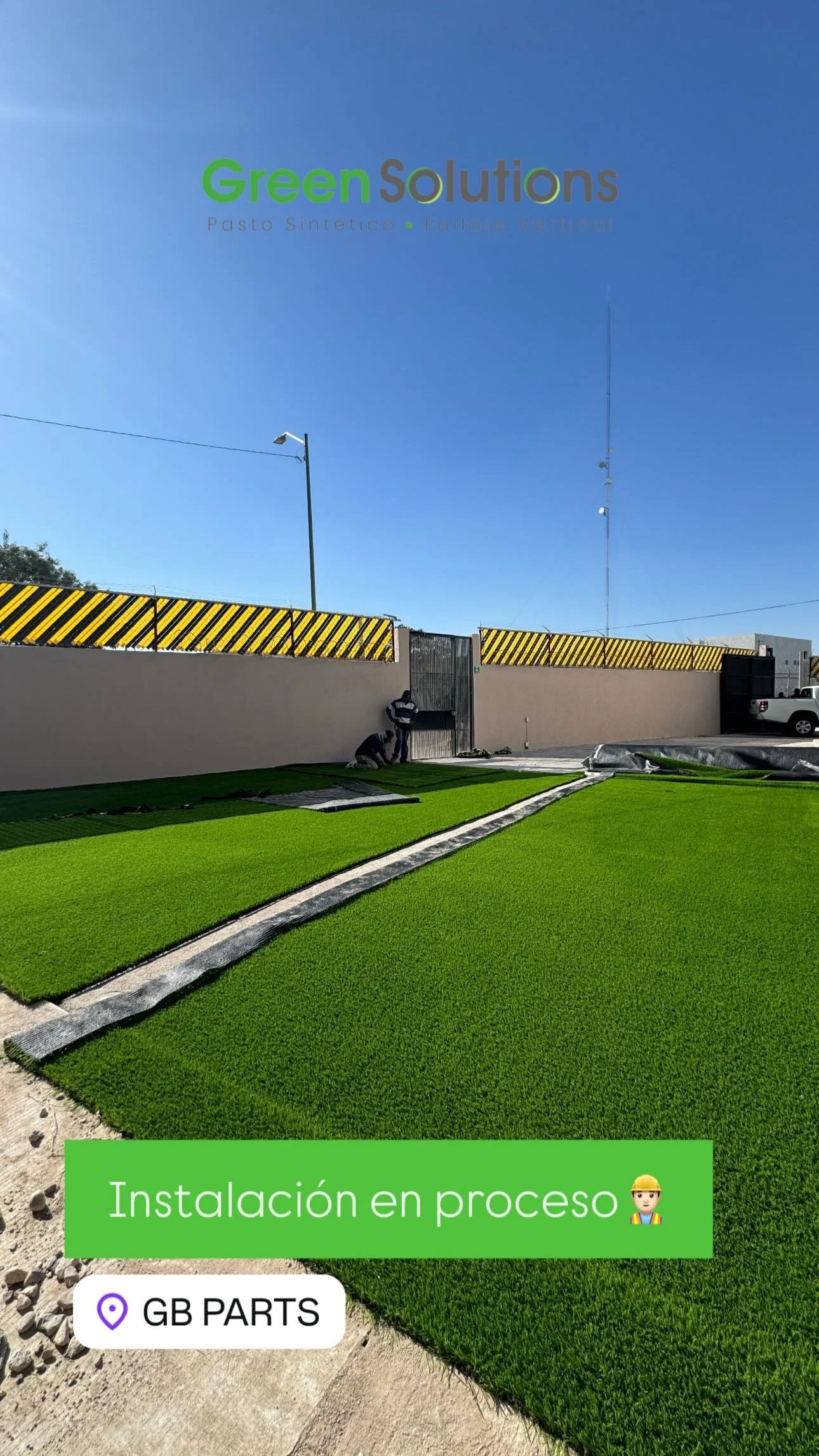 Instalación en proceso de césped artificial en un área exterior, con trabajadores asegurando las alfombrillas, cerca de una pared con aviso de advertencia, en un día soleado.