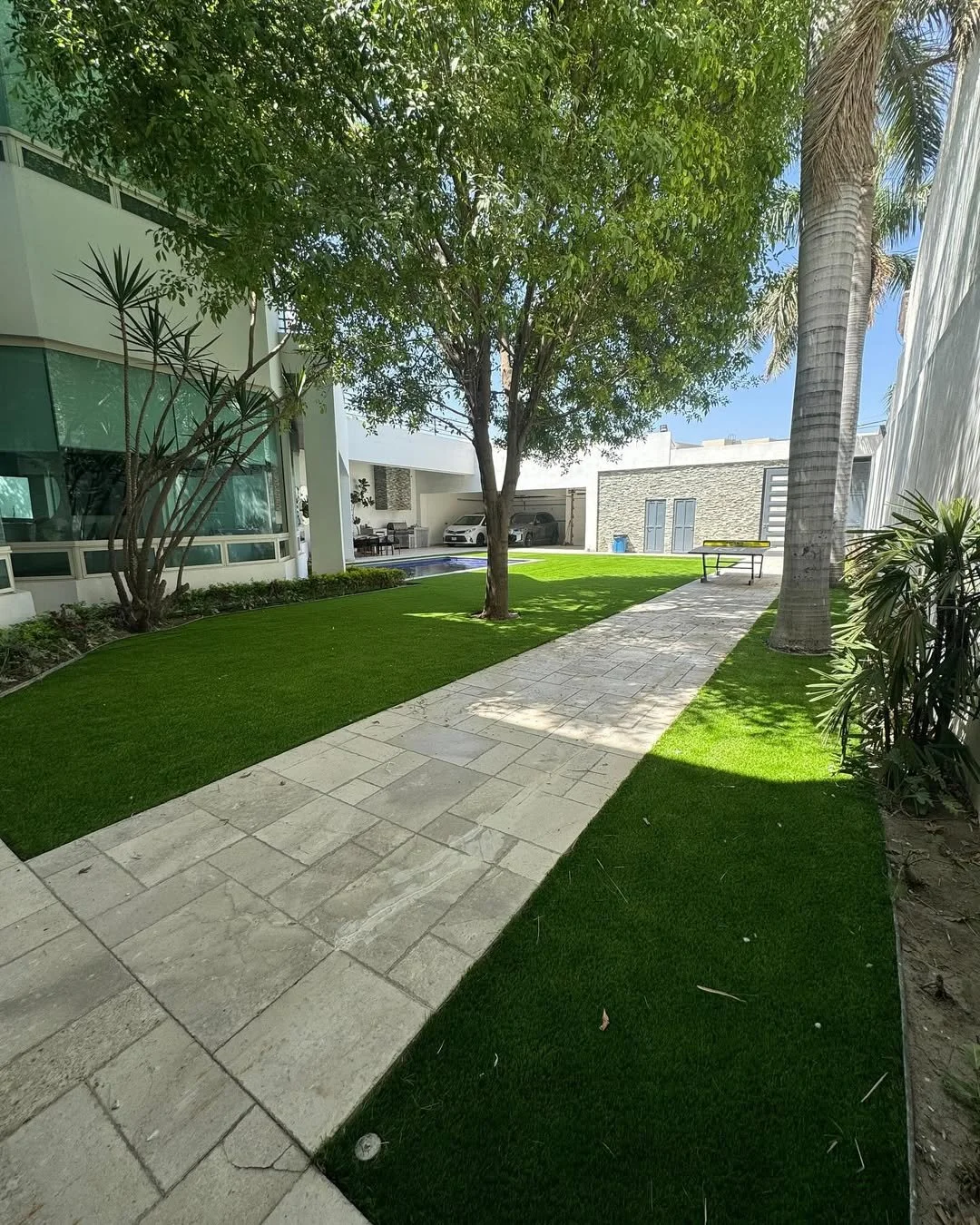 Jardín con césped verde, árboles y caminos de piedra, en un ambiente residencial moderno con casas y autos en el fondo.