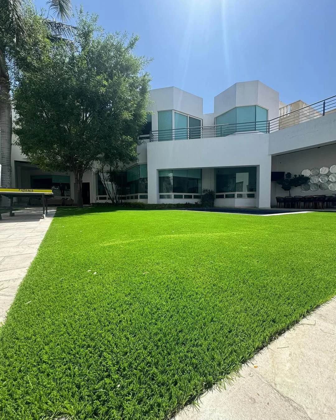 Césped verde bien cuidado en el jardín de una casa moderna blanca con muchas ventanas, árboles y cielo despejado