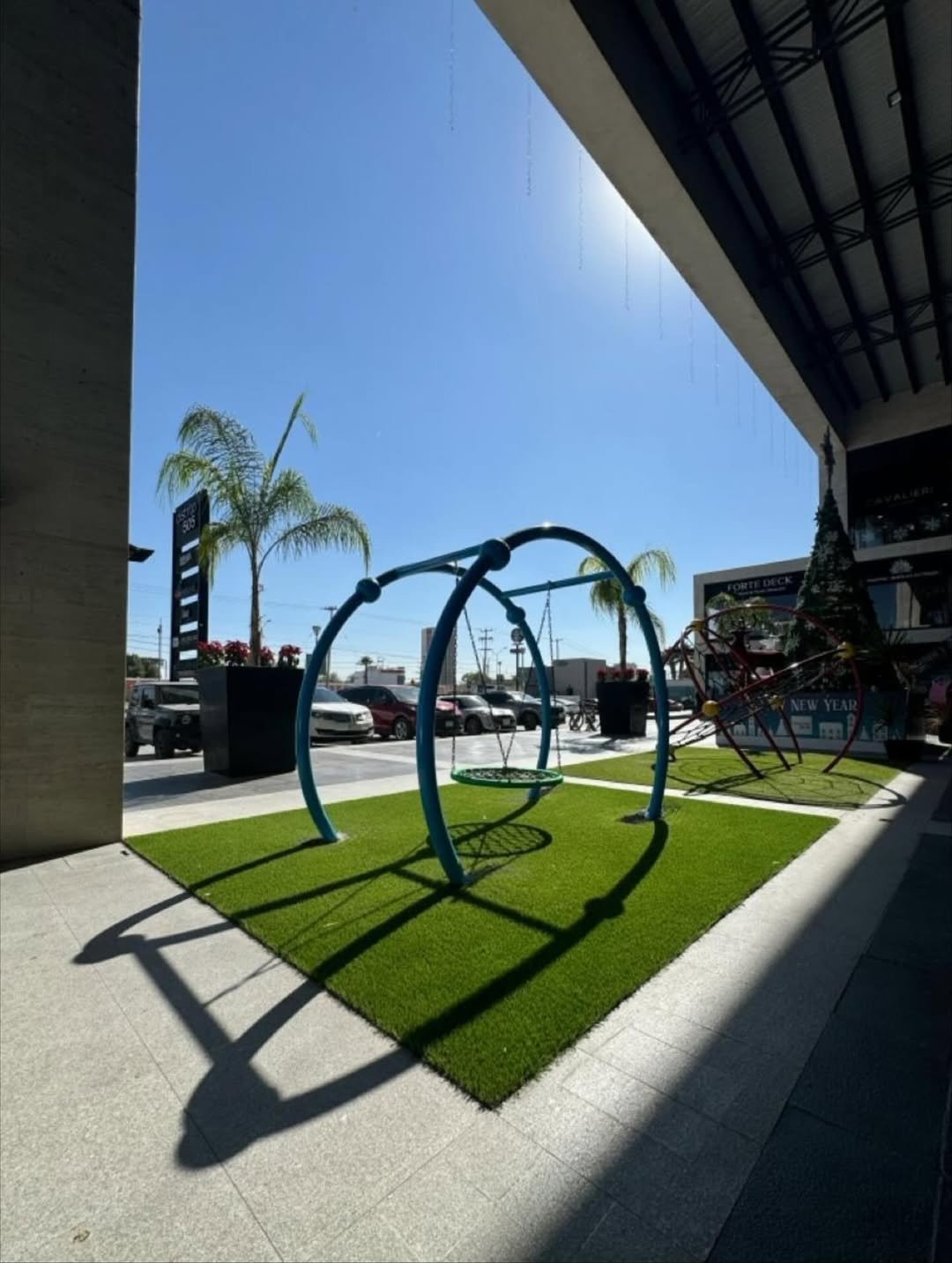 Zona Infantil con columpios en un espacio cubierto, césped artificial, árboles palmeados, y estacionamiento en el fondo bajo cielo soleado.
