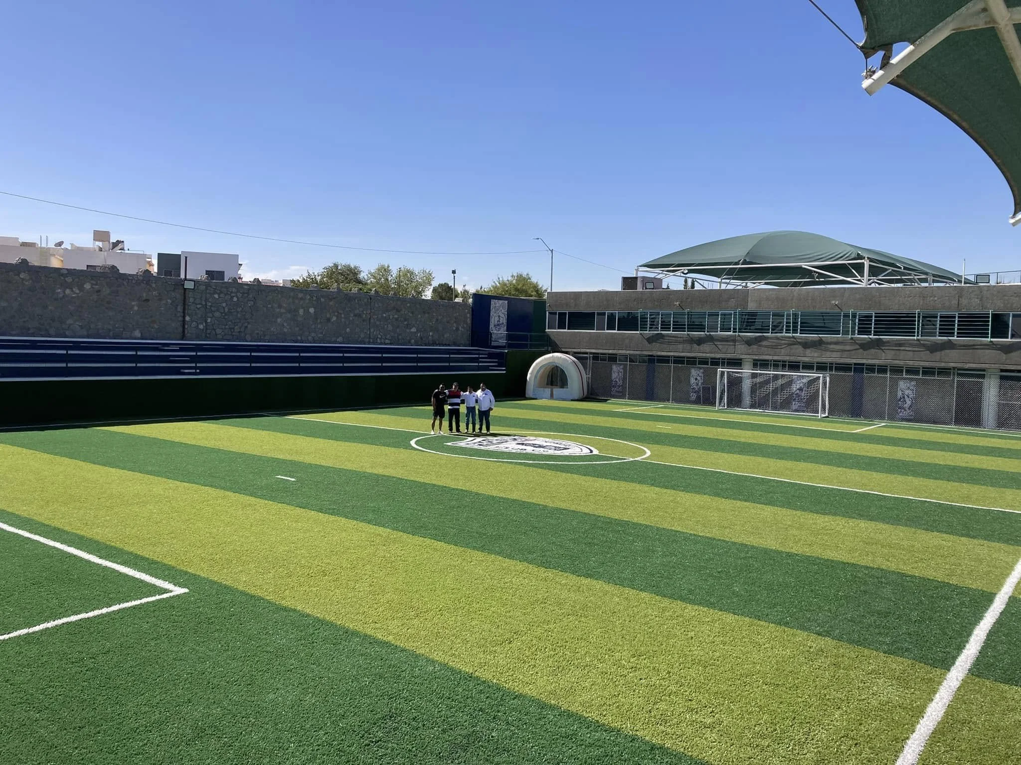 Campo de fútbol con césped artificial en un estadio con cuatro personas conversando en el centro del campo. Edificios alrededor y un cielo despejado.