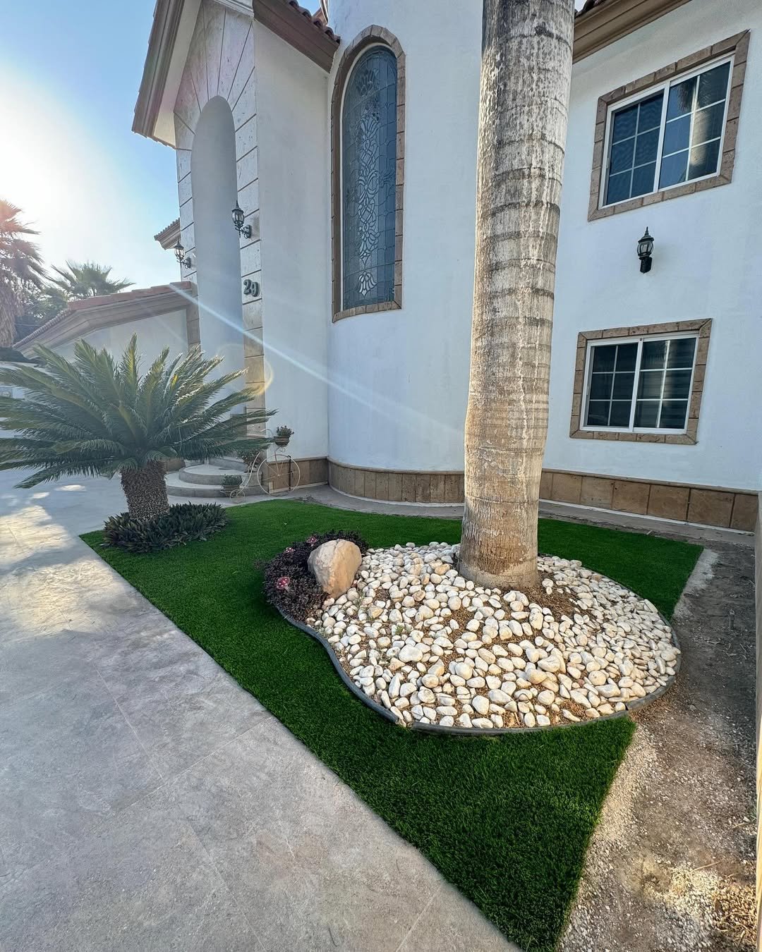 Entrada de una casa con jardín decorativo con palmera y piedras, y ventanas grandes con marcos de piedra en la fachada blanca.