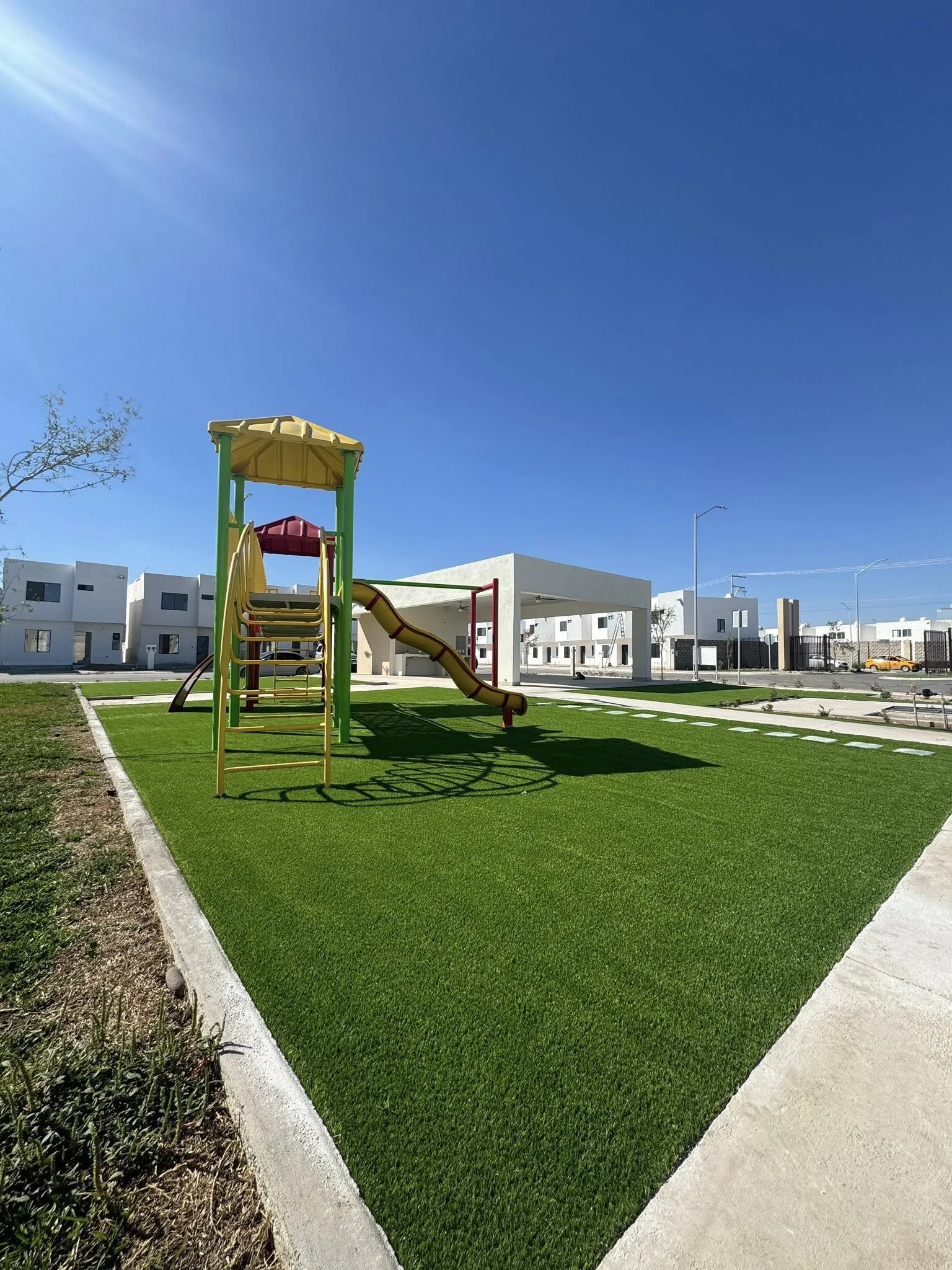 Un parque infantil con tobogán y escaleras de colores en un espacio con césped artificial, en un entorno residencial con edificios blancos y cielo despejado