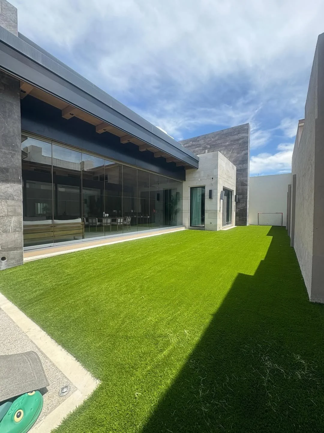 Vista de un patio exterior con césped artificial, ventanas de vidrio y paredes modernas de colores gris y beige, bajo un cielo parcialmente nublado.