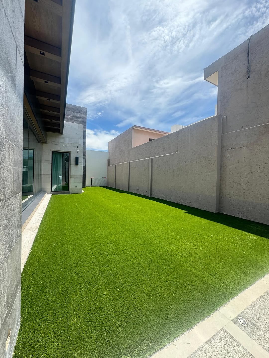 Jardín lateral con césped artificial y muro de piedra en un edificio moderno, cielo parcialmente nublado.