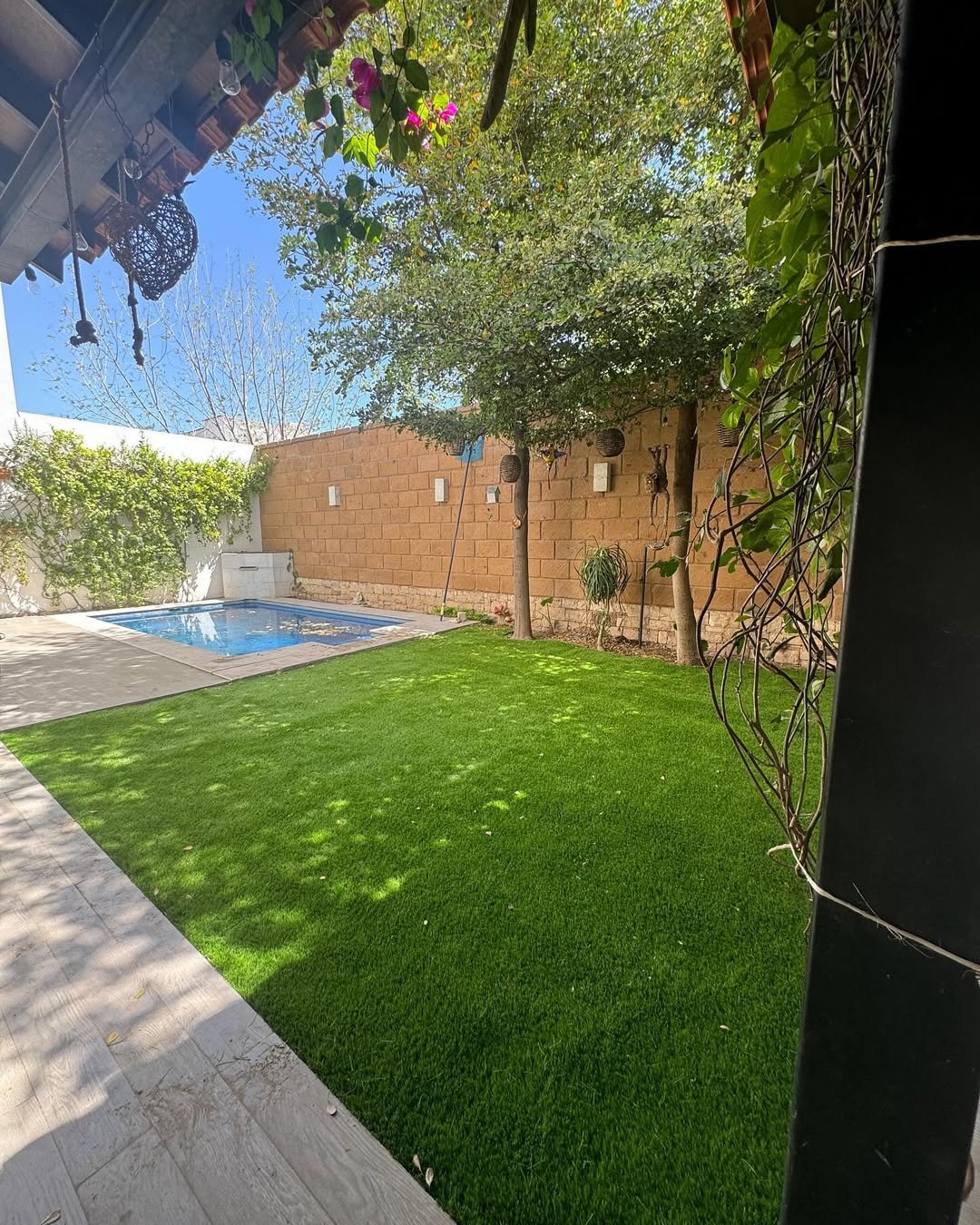 Jardín con césped verde, árboles y una piscina pequeña en el fondo, rodeado de pared de ladrillos y algunos adornos colgados.