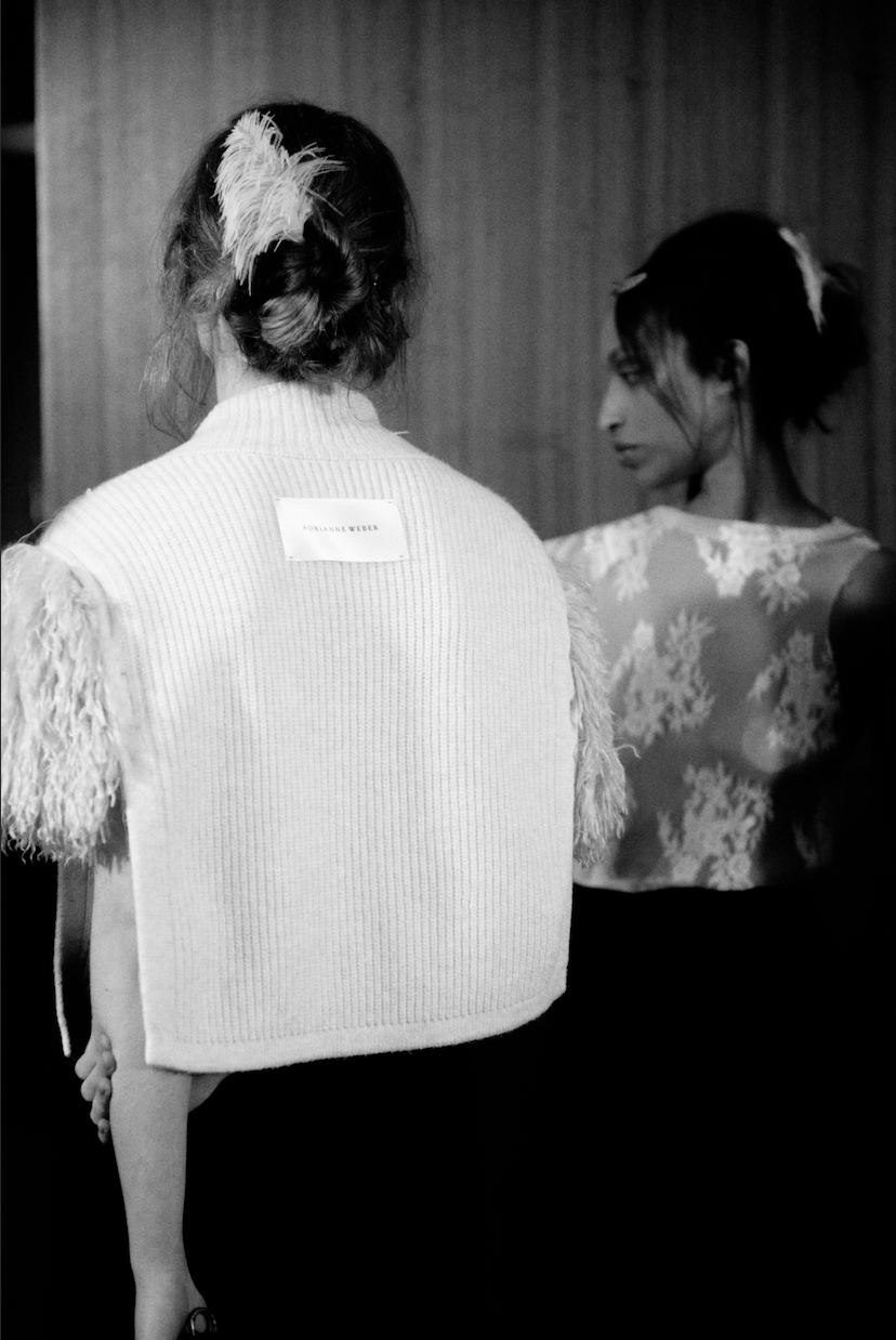 Two women with styled hair, one with a feathered hair accessory. The woman in the foreground has a cardboard label on her back that reads "Adrianna Weber." The woman in the background is wearing a floral patterned dress or top. The image is in black and white.