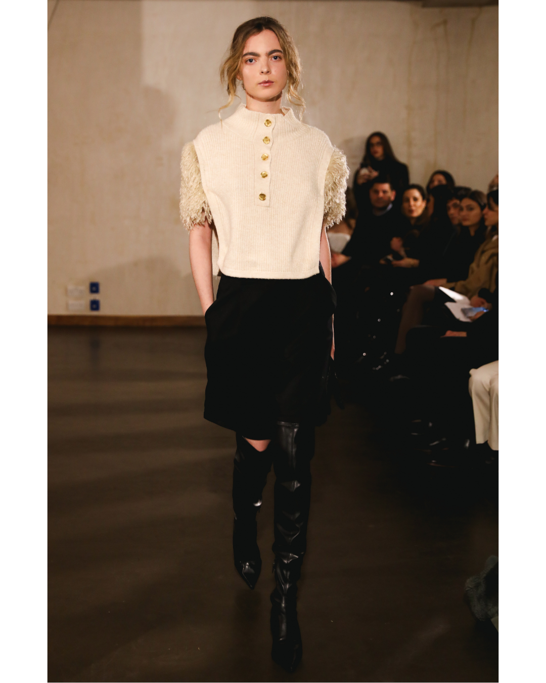 A female model walking on runway wearing a cream sweater with gold buttons and detail on the shoulders, paired with black pants and over-the-knee black boots, with audience seated on the side.