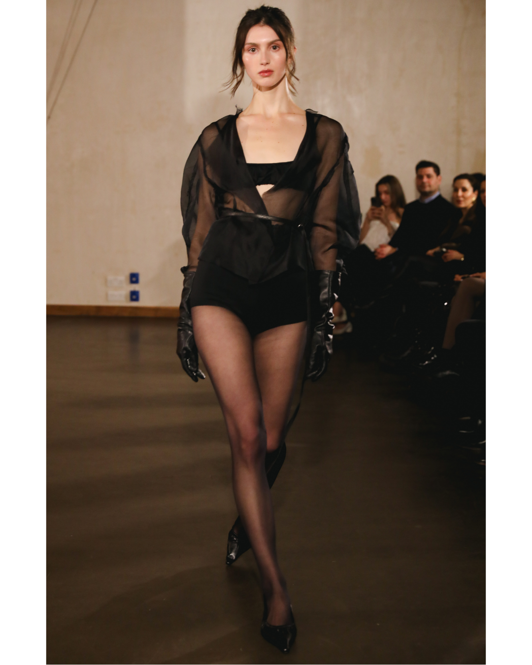 A female model walking down a runway wearing black sheer lingerie with long black gloves and high heels, during a fashion show with an audience seated on either side.