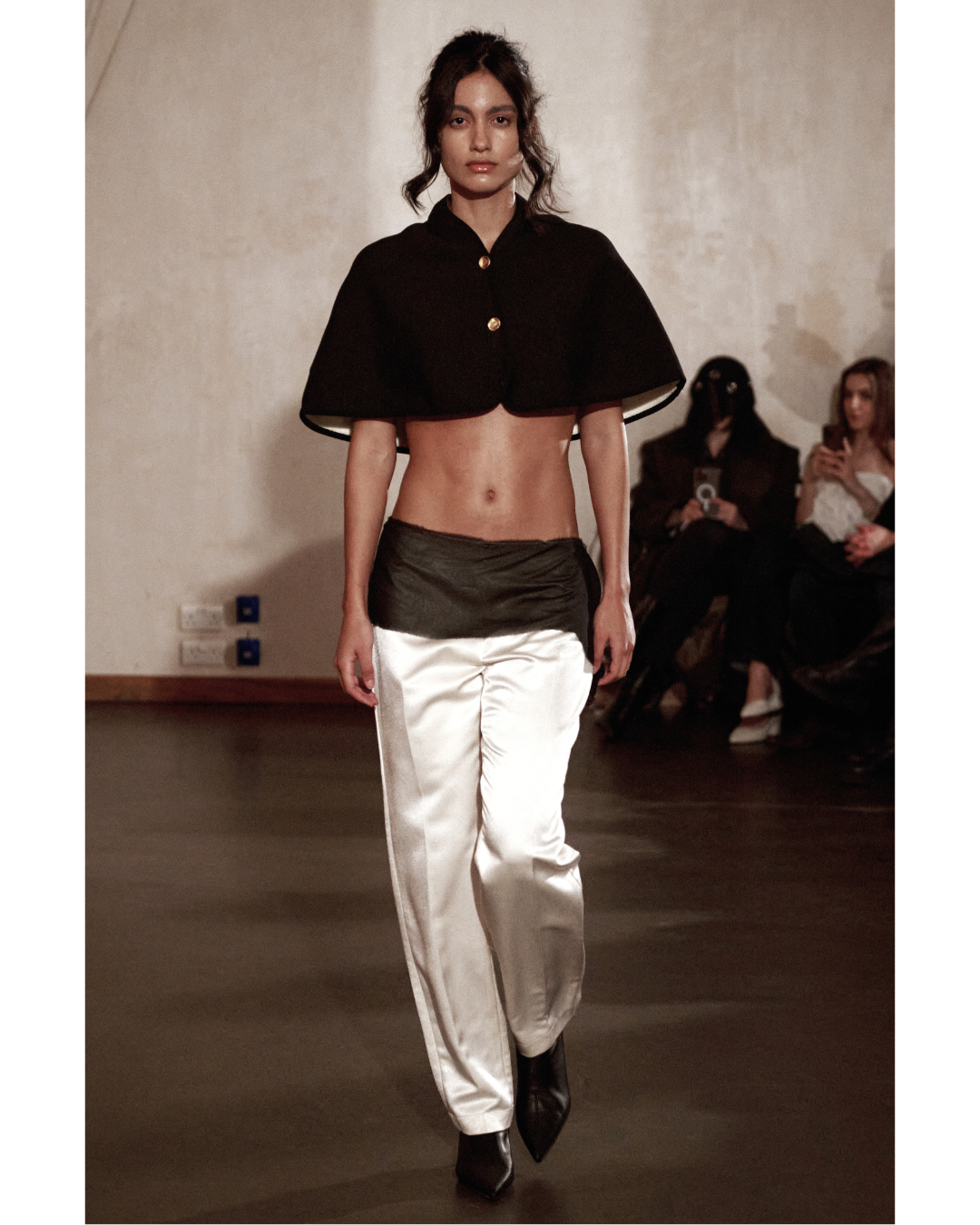 Female model walking on runway wearing a black cropped cape with gold buttons, satin loose-fitting pants, and black pointed shoes, with audience seated in the background.