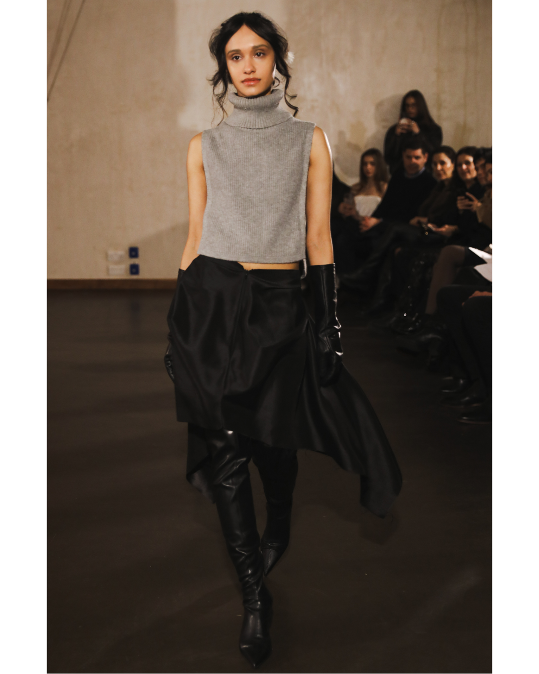 Fashion model walking on runway wearing a sleeveless gray turtleneck top, black skirt, and black thigh-high boots at a fashion show.