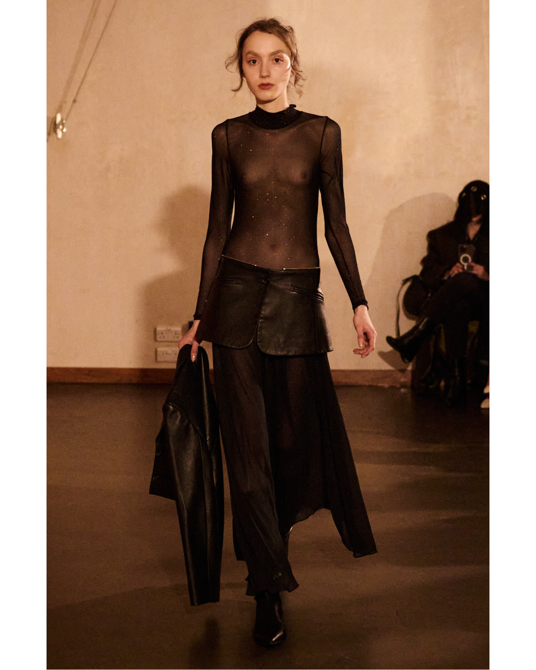 A model walking in a fashion show, wearing a sheer black long-sleeve top, a leather skirt, and black sheer wide-leg pants. She is carrying a black jacket in her left hand and has an elegant, confident expression.