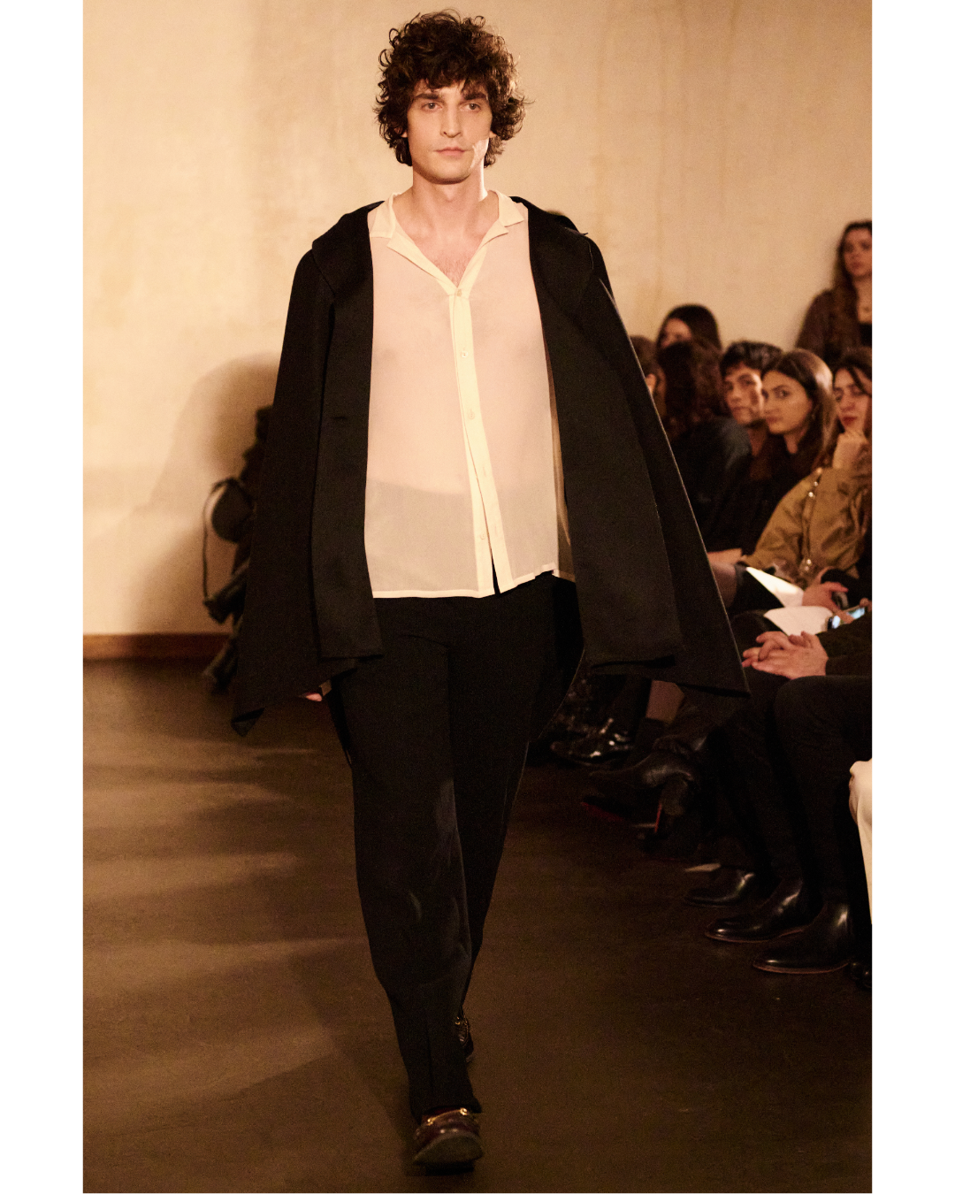 A male model with curly hair walking on a runway wearing a sheer white blouse, black pants, and a black jacket draped over his shoulders during a fashion show.