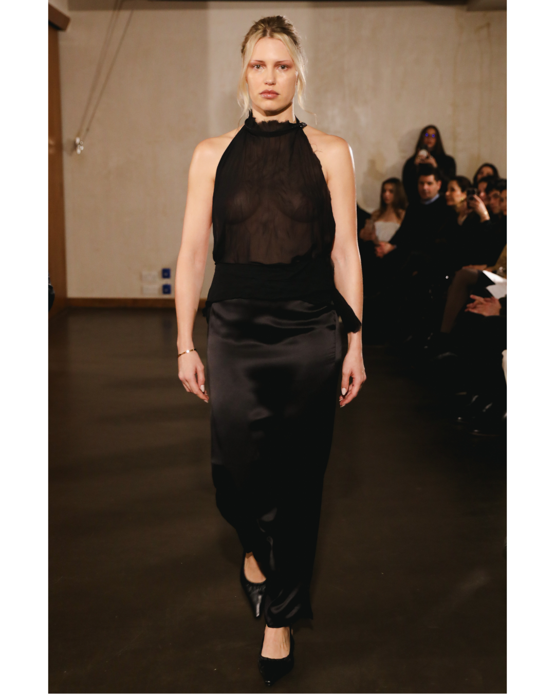 Model walking on runway wearing a sheer black sleeveless top and satin black skirt with black high heels at a fashion show.