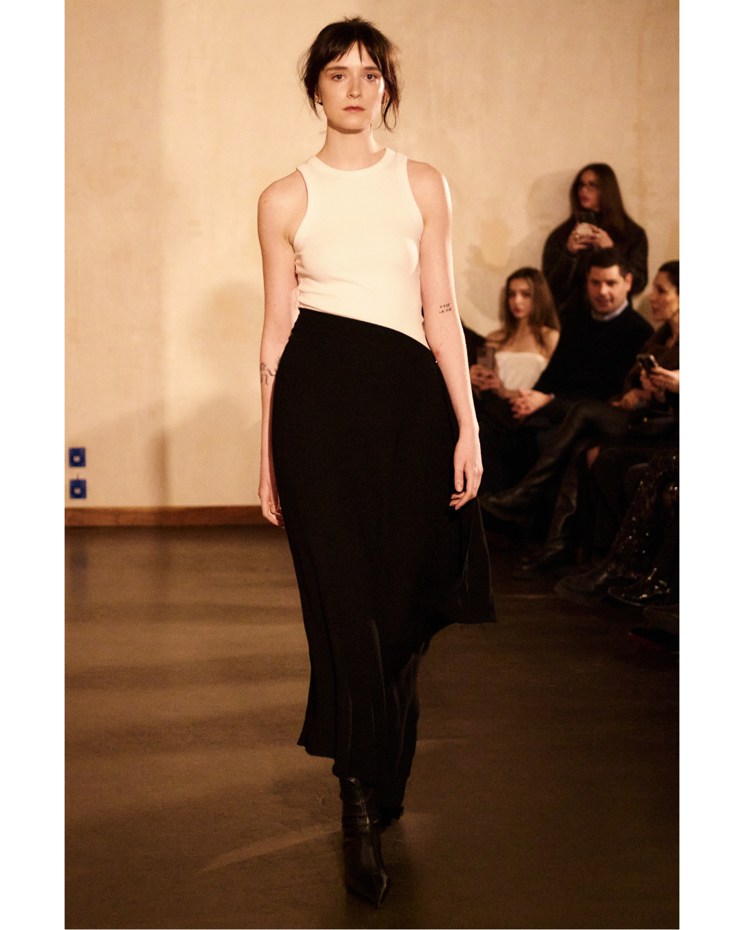 A female model walking on a runway wearing a sleeveless white top and a long black skirt during a fashion show. Audience members are seen watching and taking photos in the background.
