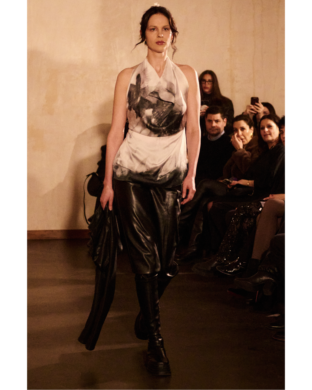 Model walking down runway wearing black leather pants and a sleeveless top with abstract black and white print during a fashion show.