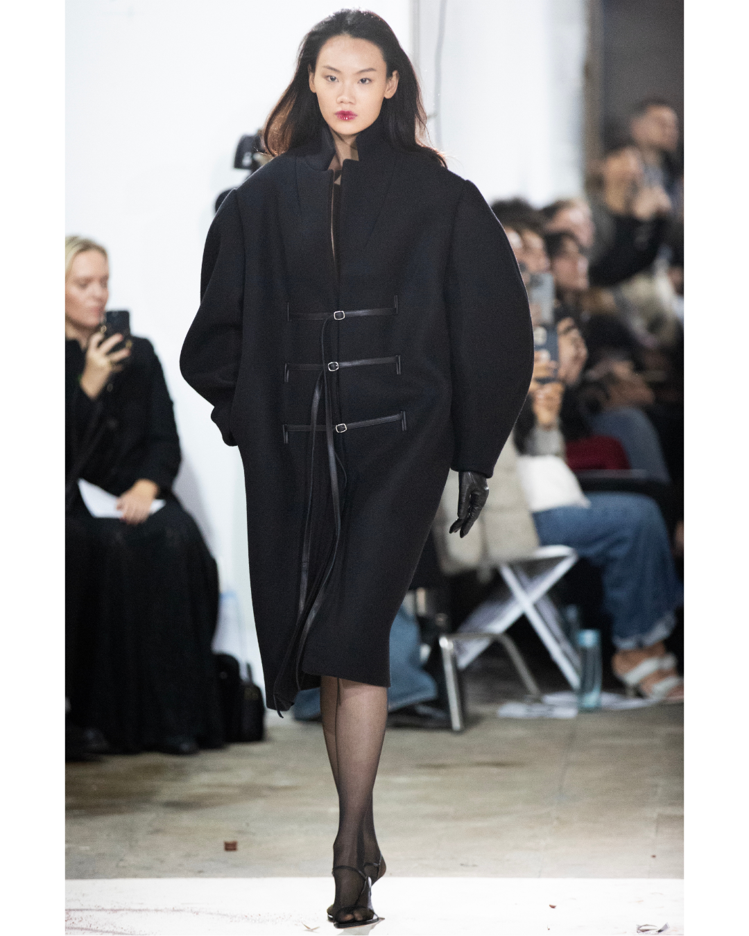 A woman walks on a runway during a fashion show, wearing a black coat, black gloves, sheer black stockings, and high heels, with spectators watching and taking photos in the background.