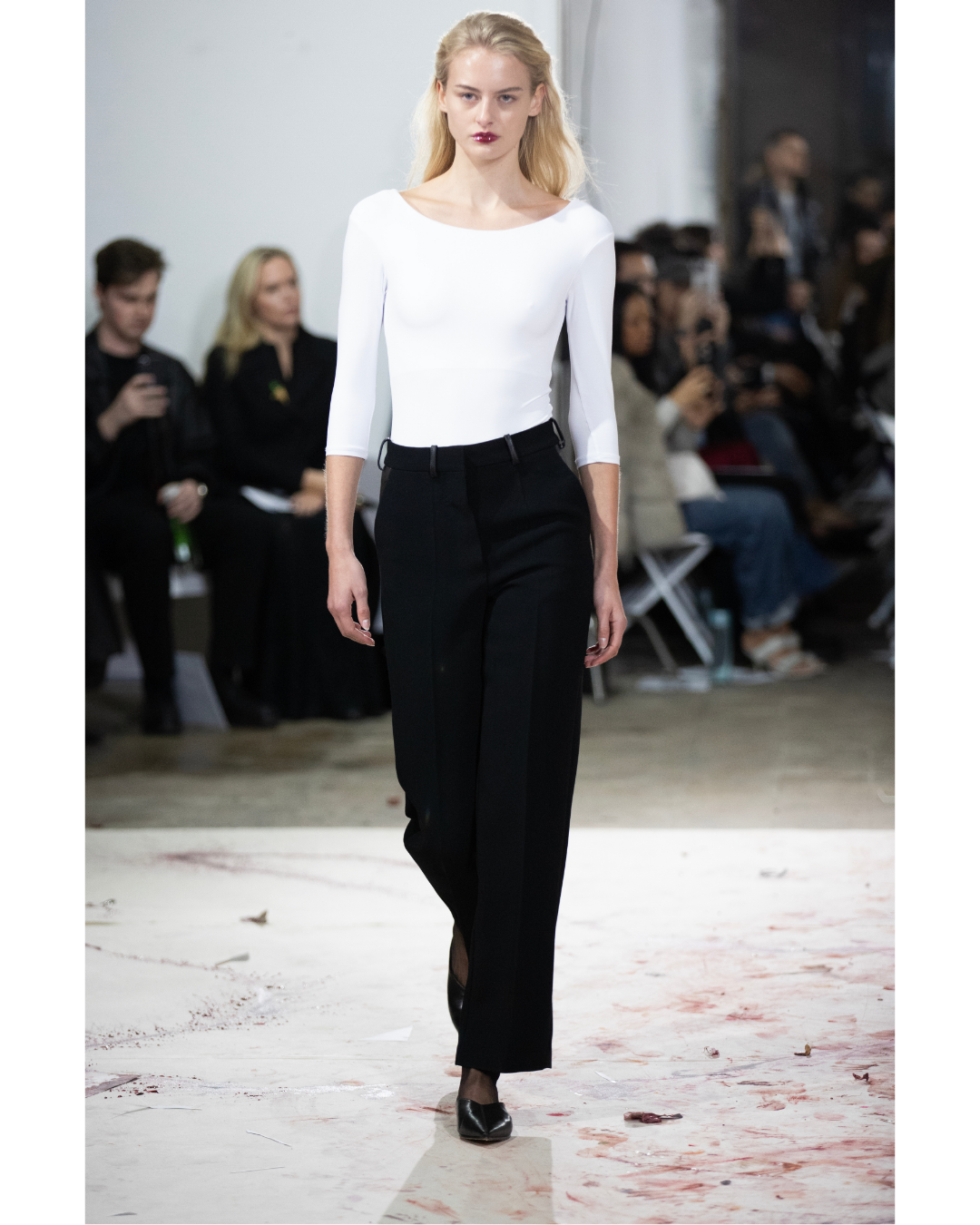 A female model walks down a runway at a fashion show, wearing a white three-quarter sleeve top and high-waisted black trousers, with a blurred audience in the background.
