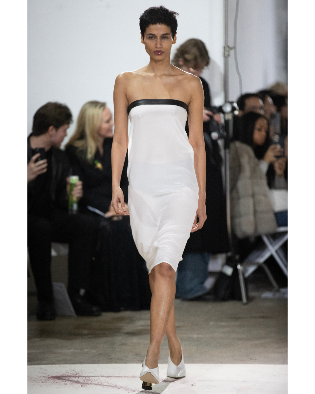 Model walking down the runway wearing a strapless, white dress with black trim, at a fashion show with audience members in the background.
