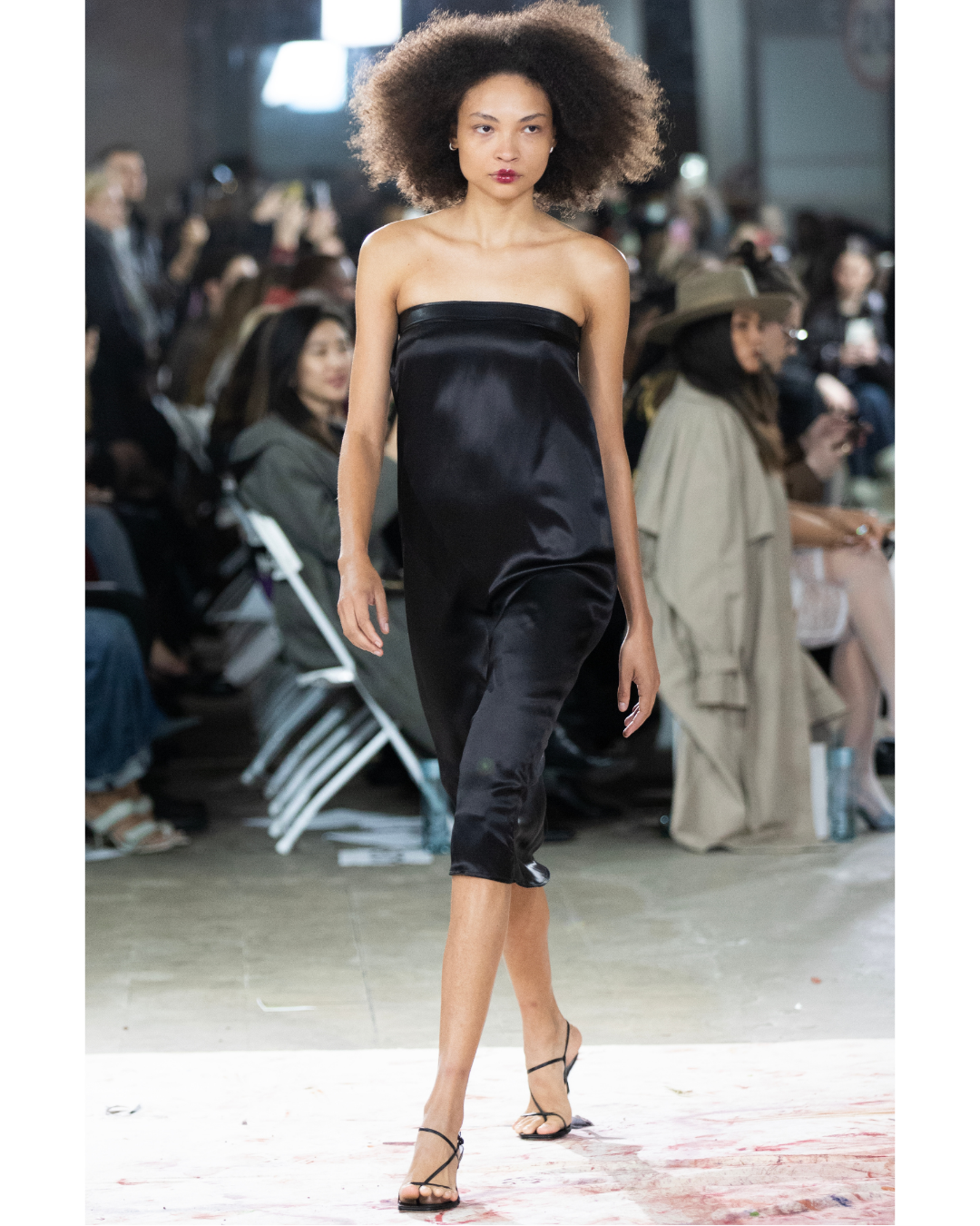 A woman walking on a runway at a fashion show, wearing a black satin strapless dress and black high-heeled sandals, with an audience seated in the background.