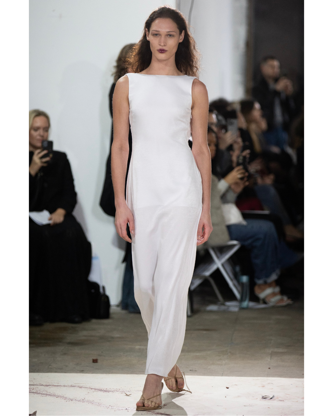 A female model walking on a runway in a sleeveless white dress with black accents on the sides, surrounded by an audience taking photos.