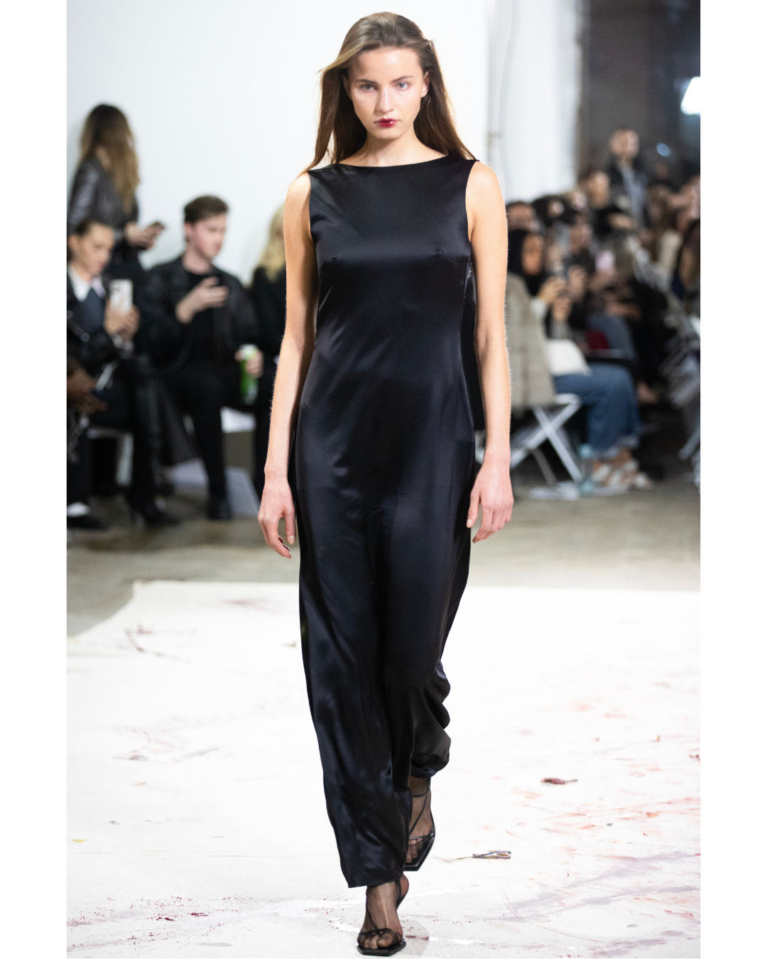 A female model walks down the runway in a sleeveless black satin dress with sheer black heels, with a fashion show audience in the background.