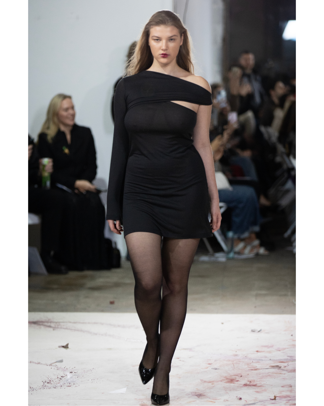 Model walking down runway wearing a black, asymmetrical dress with sheer black tights and shiny black high heels, with audience in background at fashion show.
