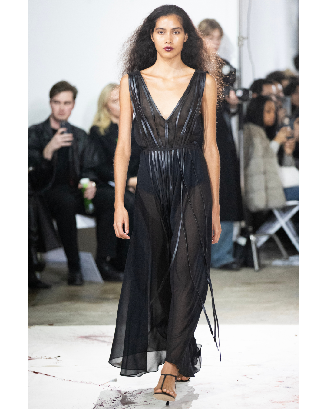 A woman walks on a runway wearing a sheer black dress with metallic accents at a fashion show.