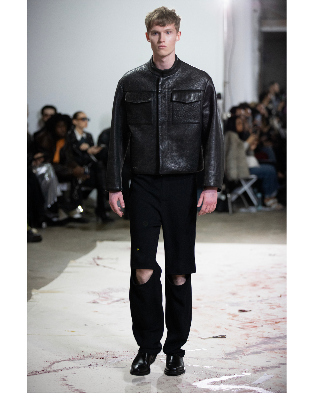Male model walking on a runway wearing a black leather jacket, black pants with a ripped knee, and black boots, with an audience watching.