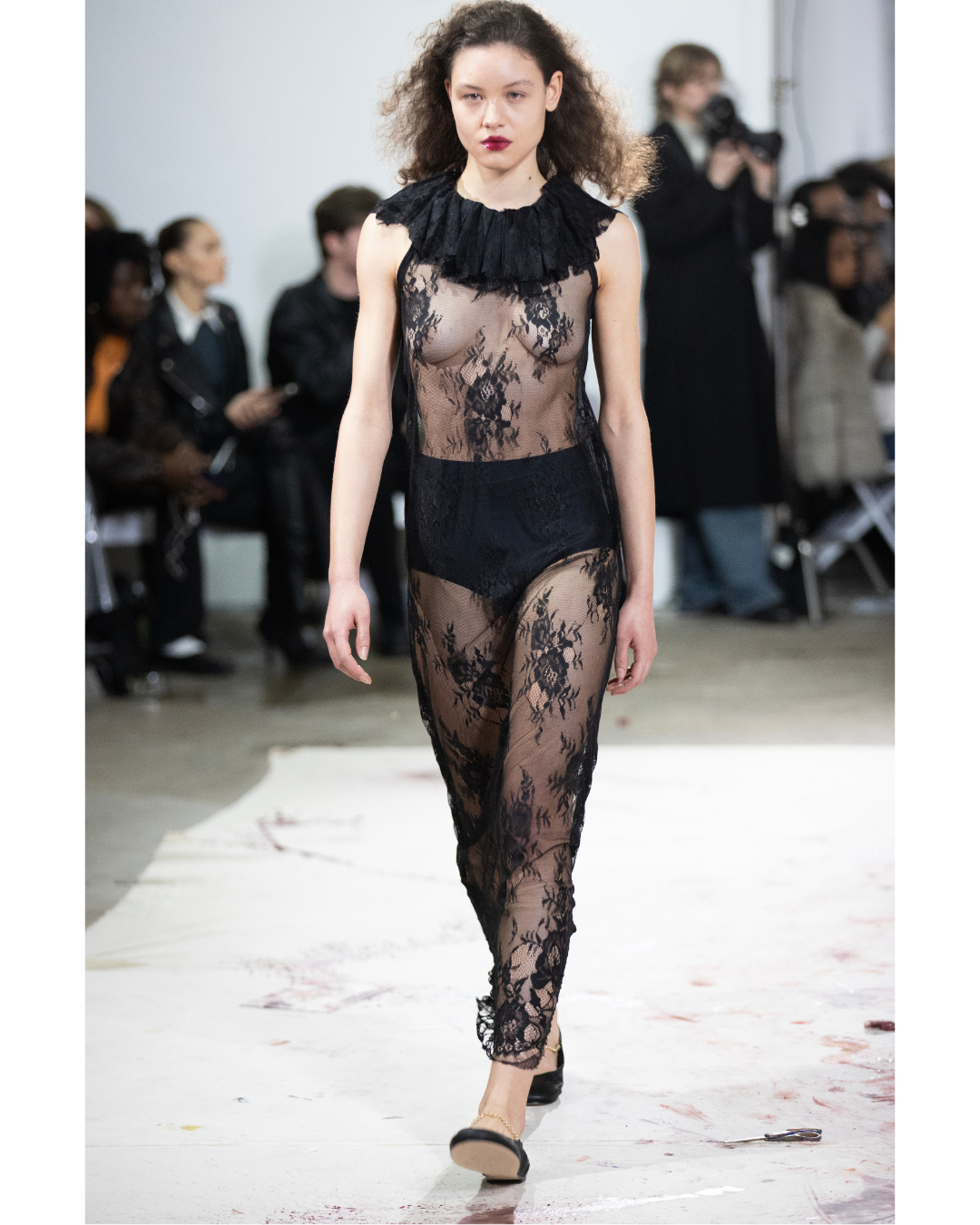 Model walking down a runway wearing a sheer black lace outfit at a fashion show.