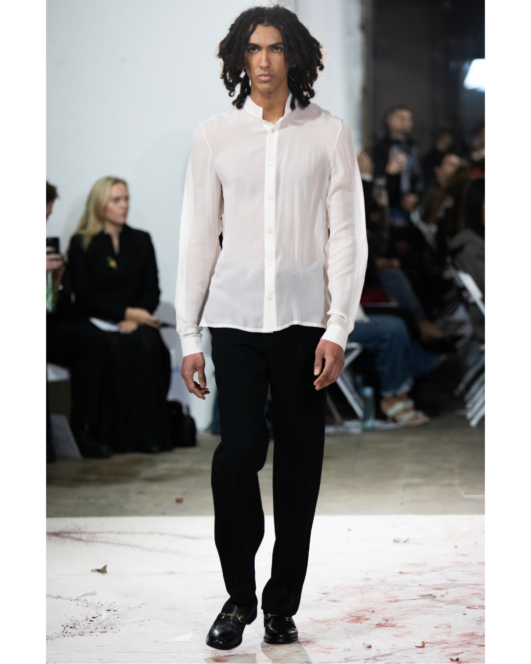 Model walking on runway in white sheer button-up shirt and black trousers at fashion show with audience seated in background.