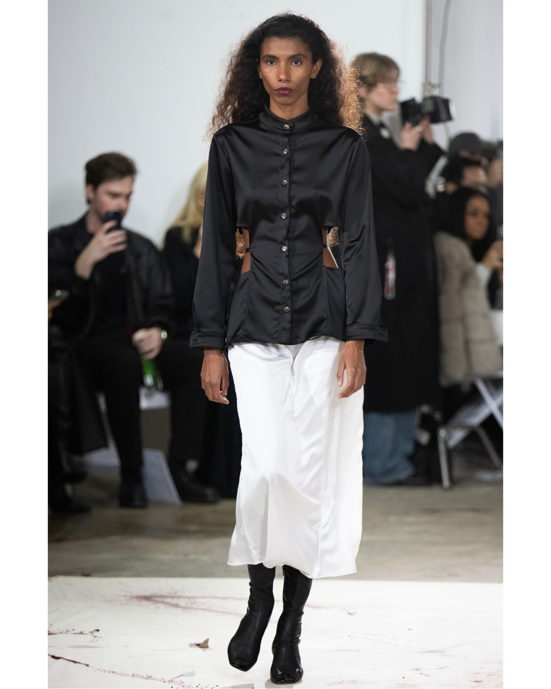Model walking on runway during fashion show, wearing black button-up shirt, white skirt, and black boots, with audience and photographers in background.