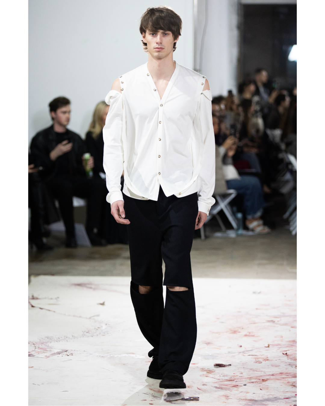 Male model walking on runway in white button-up shirt with cut-out shoulders and black ripped jeans, with audience seated in the background.