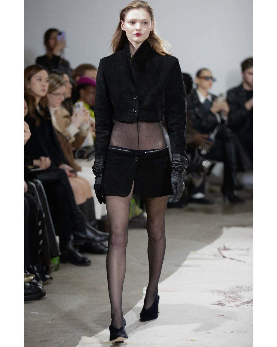 A female model walking on the runway at a fashion show, wearing a black cropped jacket, sheer top, black shorts, black gloves, sheer stockings, and black shoes, with an audience seated on either side of the runway.