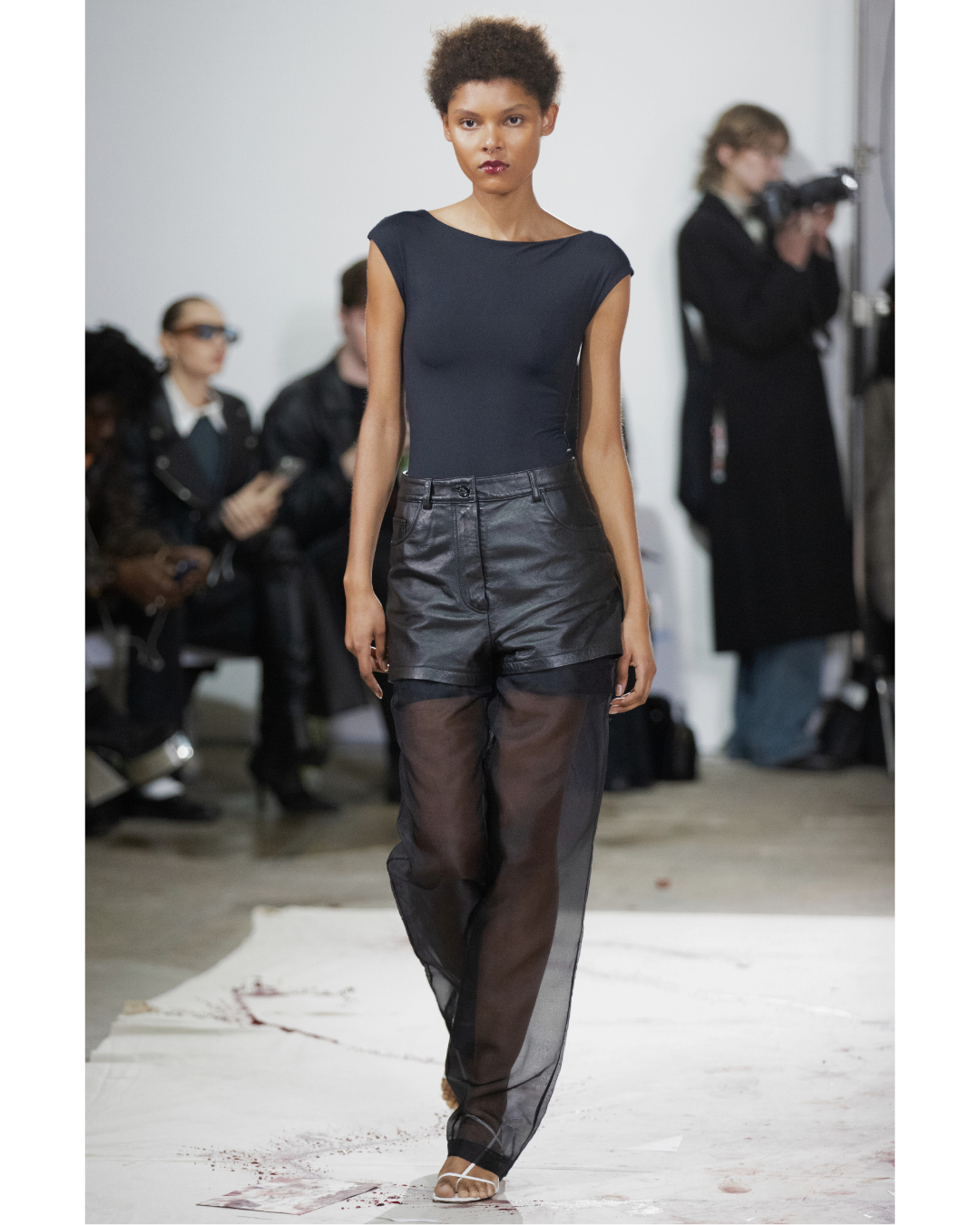 Fashion runway model wearing black top and layered pants walking on white runway with blood splatters, audience and photographers in background.