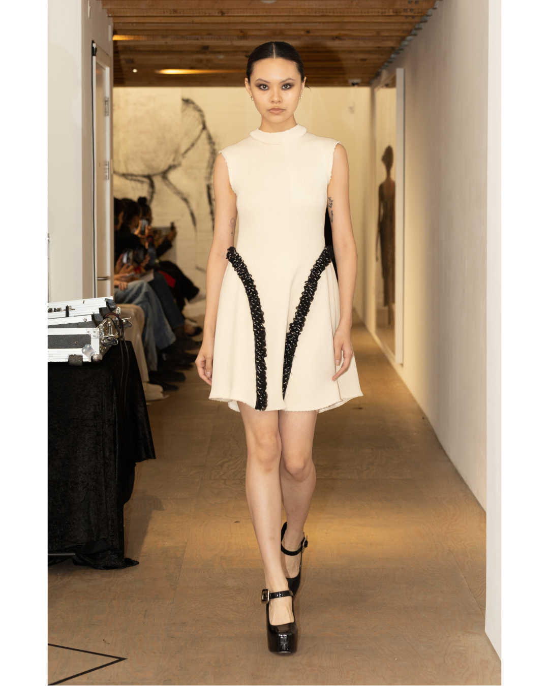 A female model walking down a runway during a fashion show, wearing a sleeveless, cream-colored dress with black decorative accents and black high-heeled shoes.