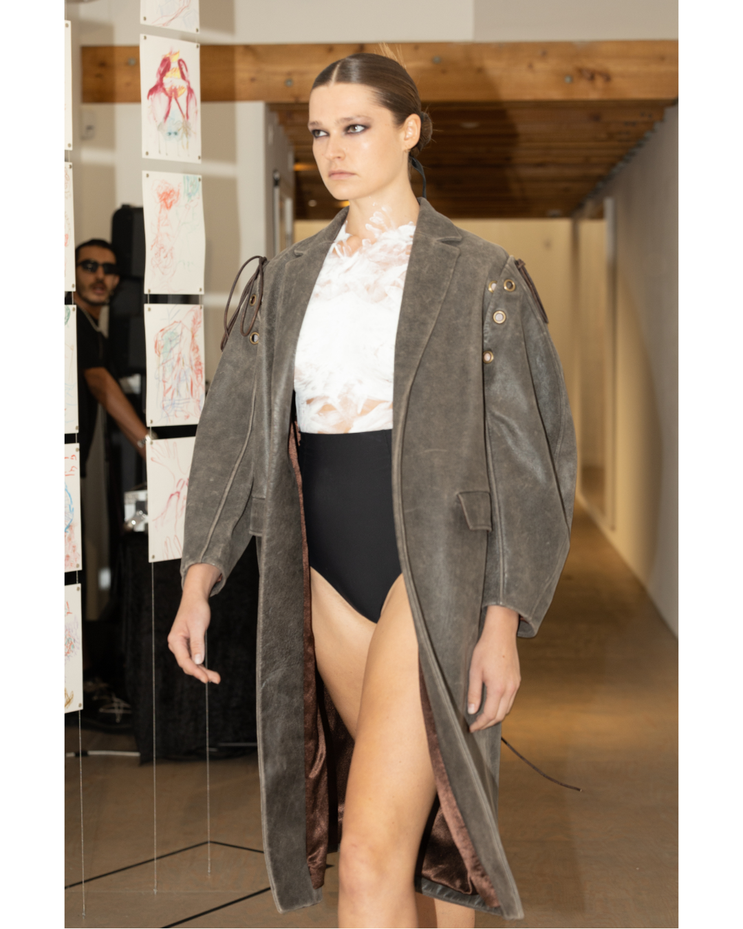 A woman models a fashion outfit with a long gray coat, a sheer white top, and black bottoms, walking down a runway.