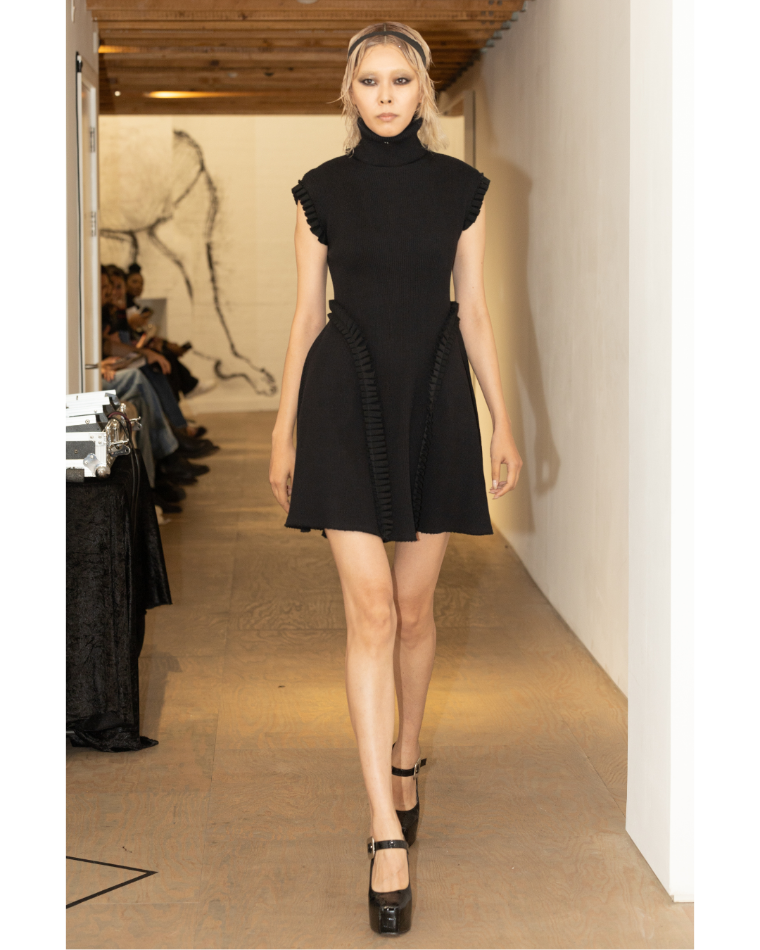 Model walking down runway during a fashion show, wearing a black dress with ruffled accents and platform heels.