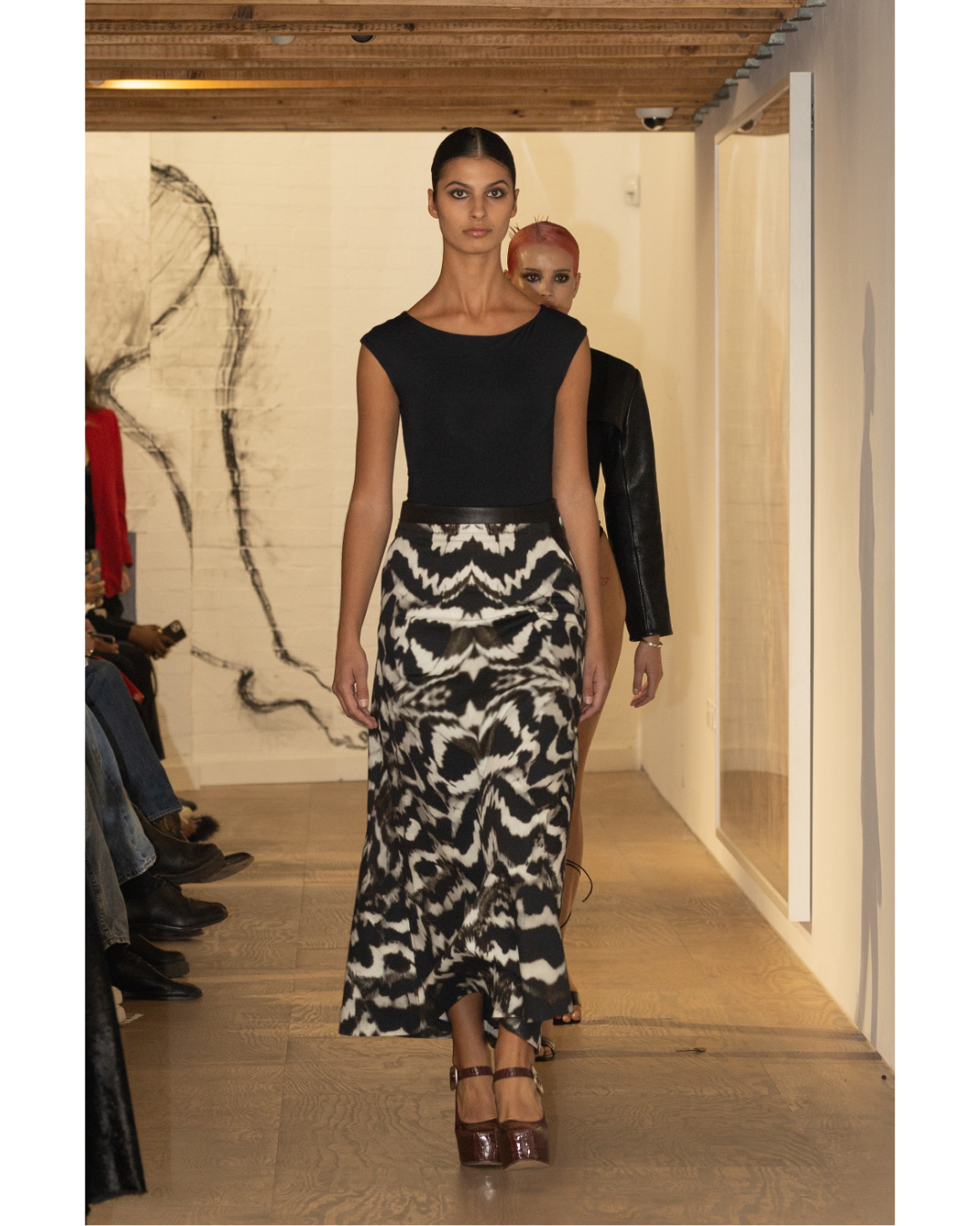 Models walking down a runway showcasing black and white patterned dresses in an indoor setting with a wooden ceiling, audience seated on the side, and artwork on the wall.