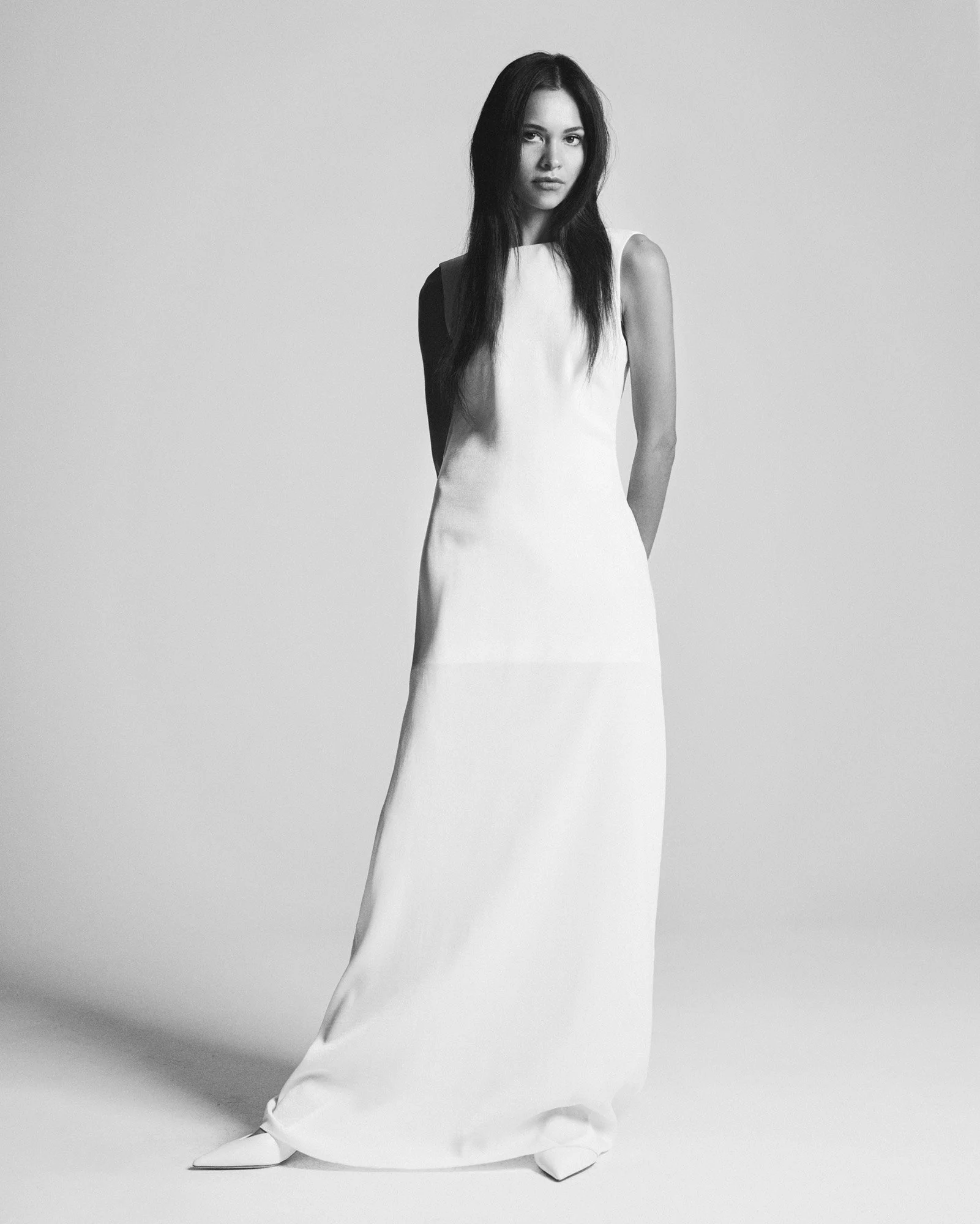 A woman in a long, sleeveless white dress standing on a plain background.