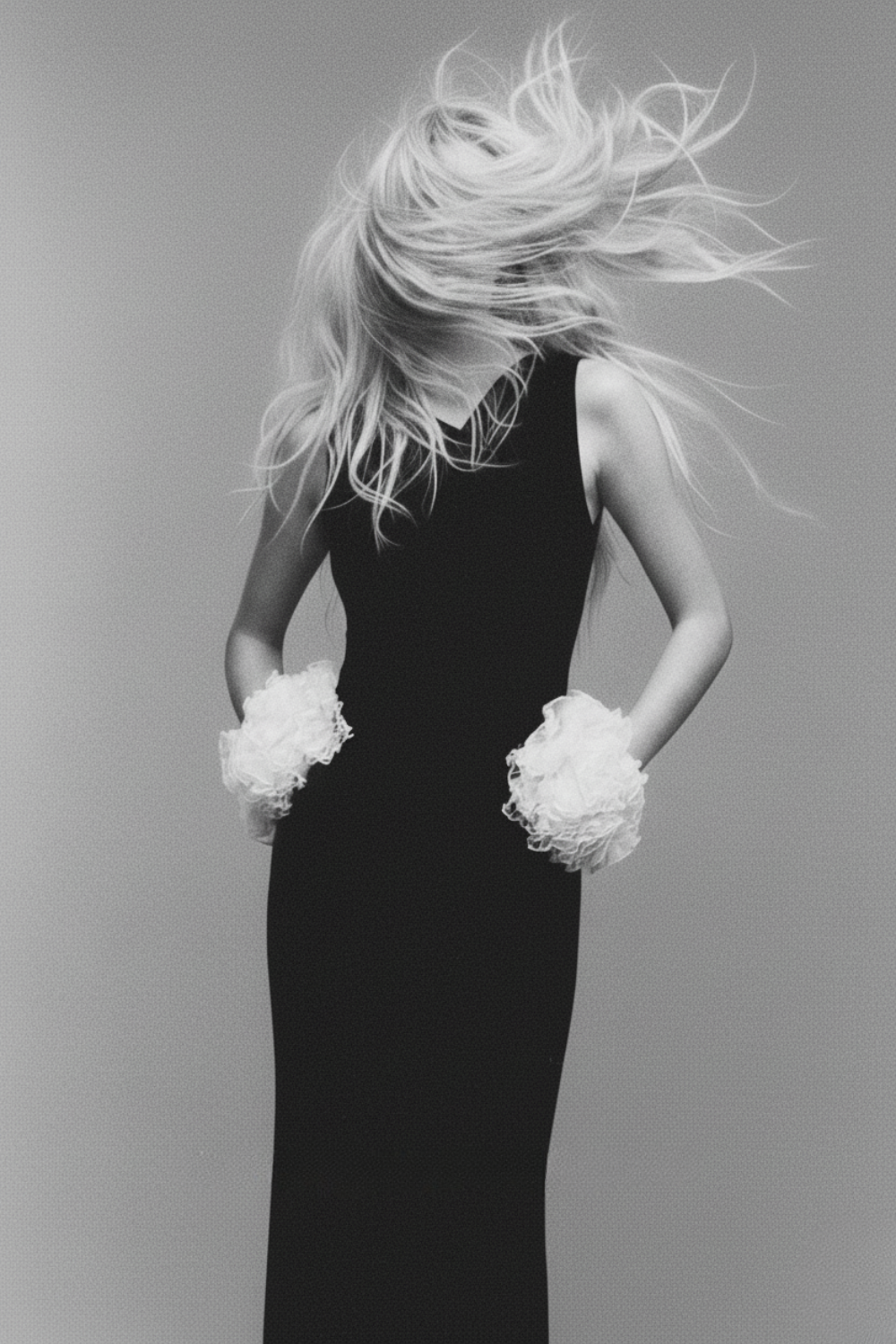 A woman in a long black dress with fluffy wrist cuffs, her hair flying around her face, holding her hands in her pockets, against a simple plain background.