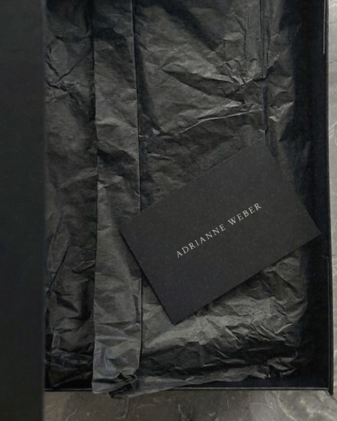 Black gift box with crumpled black tissue paper and a black card that has the name "Adrianne Weber" printed on it.