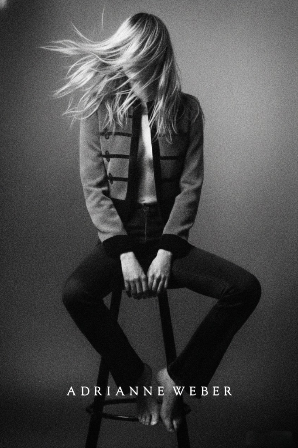 A black and white photo of a woman with long hair, turned away, hair blowing, sitting on a stool with legs spread, wearing a jacket and pants, with the name Adrianne Weber written at the bottom.