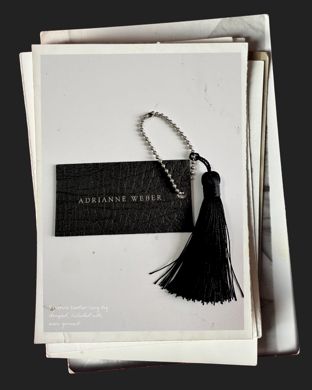 A signature leather hang tag with a pearl and black beaded chain and black tassel, placed on top of a white card with the name 'Adrianne Weber' printed on it. The card is on a stack of other white papers.