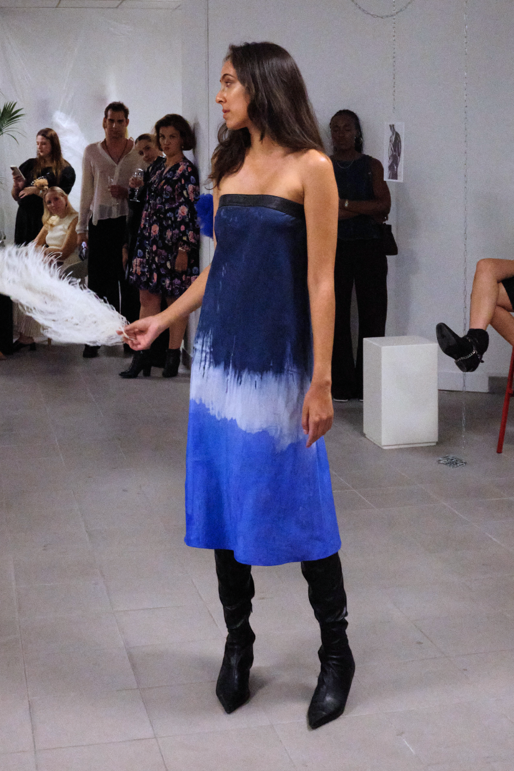 Woman in a strapless blue ombré dress and black boots holding a white feather at a fashion event with people in the background.